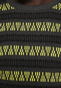 Pull en tricot présentant un motif géométrique de rayures jaunes et noires. La texture est lisse avec un col rond. Les contrastes de couleurs mettent en valeur le design.