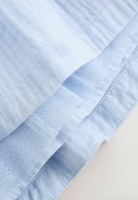 Helles blaues Gewebe mit einer strukturierten Oberfläche, das eine Kombination aus glatten und netzartigen Abschnitten aufweist, die schichtweise übereinander liegen.