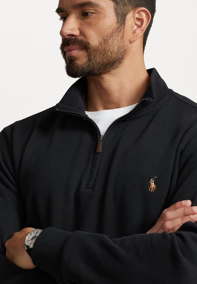 Black zip-up sweater made of a textured knit fabric. Features a small logo on the chest and a brown zip pull detail. White shirt underneath.