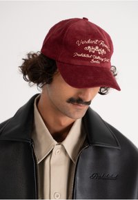 Burgundi värvi ribakanga müts, mille esipaneelil on tikand "Verdant Reserve Prohibited Clothing Group Berlin"; kumer visor, tekstuuriga kangas.