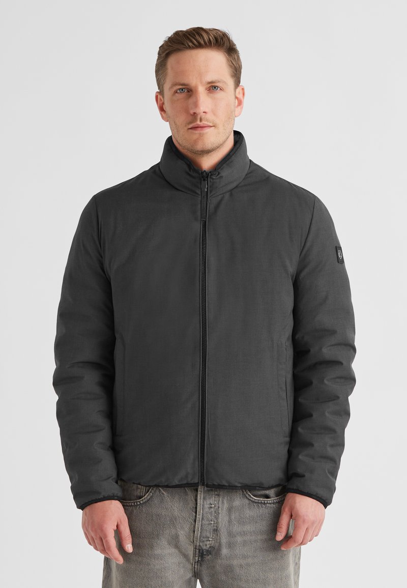 Schwarze, aufziehbare Jacke mit hohem Kragen, gesteppter Textur und taillierten Ärmeln. Verfügt über ein Logo am linken Arm. Getragen über grauen Jeans.