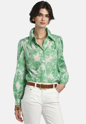 Frau trägt eine grüne, durchsichtige Bluse mit floralem Muster und aufgepufften Ärmeln, weiße Hose, braunen Gürtel mit goldener Schnalle und goldenen Schmuck, posiert mit einer Hand in der Tasche.