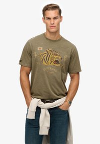 Camiseta de algodón verde olivo con un gráfico de tigre, la bandera japonesa y texto. Usada con jeans azules y un sudadera blanca atada a la cintura.