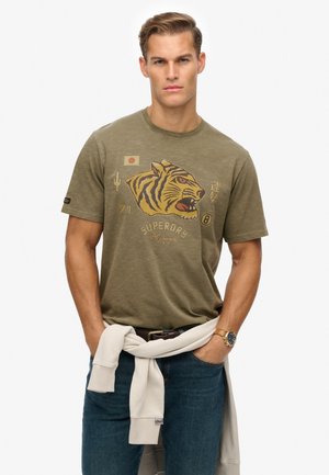 Olive green cotton t-shirt featuring a tiger graphic, Japanese flag, and text. Worn with blue jeans and a white sweatshirt tied at the waist.
