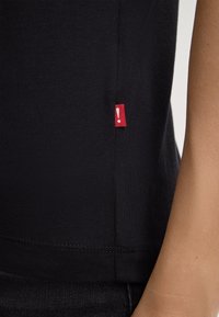 T-shirt en coton noir avec une étiquette en tissu rouge comportant un détail en forme de point d'exclamation sur le côté gauche près de l'ourlet. Texture lisse.