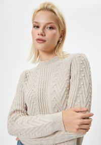 Jasnobeżowy sweter dzianinowy z falbaniastym dekoltem, teksturowanymi wzorami w zygzak oraz kulkami na rękawach.