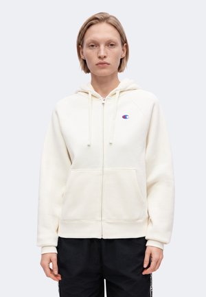 Persoon draagt een witte hoodie met rits met een blauw en rood logo op de borst en zwarte broek, staand tegen een effen lichte achtergrond.