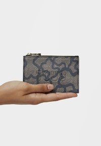 Textured wallet in beige with a dark blue abstract pattern, featuring a zip closure and held in a hand. Accents in gold-tone hardware.
