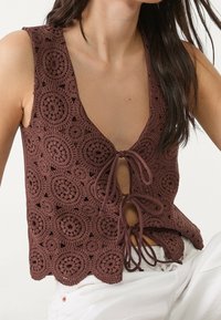 Brązowy top szydełkowy z głębokim dekoltem w kształcie litery V, z okrągłymi wzorami, zawiązywany z przodu i z falbaniastym wykończeniem krawędzi.