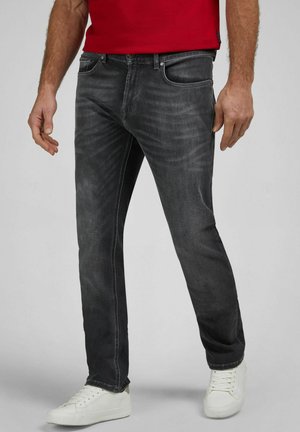 Man draagt versleten zwarte spijkerbroek, witte sneakers en een rood shirt, staande tegen een eenvoudige grijze achtergrond.