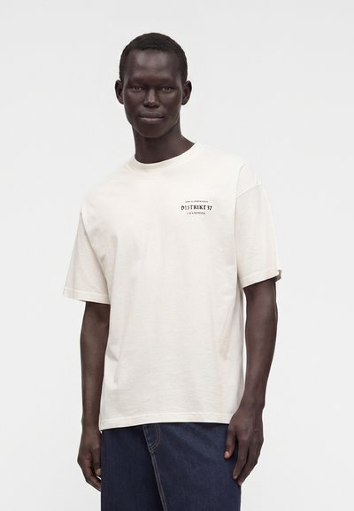 Άνδρας με λευκό φαρδύ T-shirt με μικρό μαύρο κείμενο στο στήθος και σκούρο μπλε τζιν, σε απλό ανοιχτό φόντο.