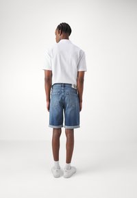 Vitt kortärmad skjorta, ljusa denimshorts med uppvikt fåll, och vita sneakers med färgade accenter, sedd bakifrån.