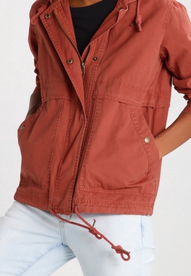 Personne portant une veste zippée couleur rouille avec cordons et poches à boutons-pression, associée à un jean bleu clair. Mains dans les poches de la veste.