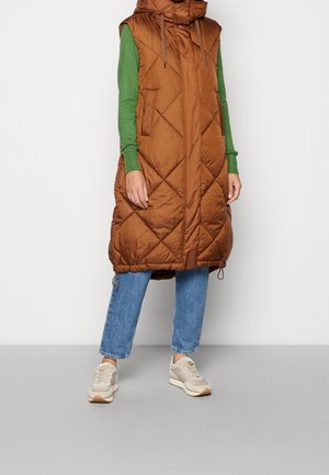 Personne portant une longue veste matelassée marron sans manches par-dessus un haut vert à manches longues, un jean bleu et des baskets beige, debout à l'intérieur.