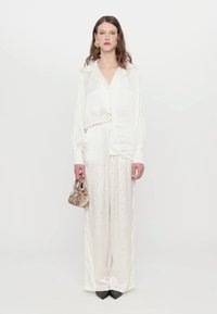 White satin blouse with a collared design, paired with loose white pants. Holding a small patterned handbag, black pointed shoes visible.