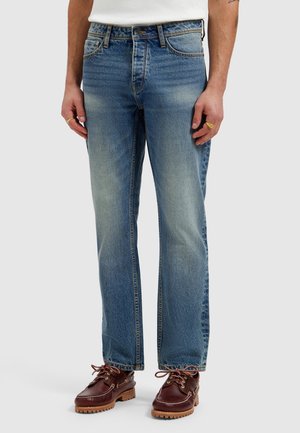 Man wearing blue faded straight-leg jeans, white shirt tucked in, brown leather lace-up shoes, and gold rings on left hand.