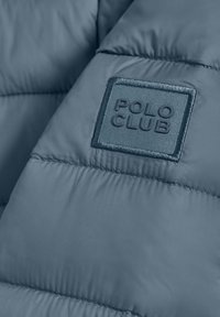 Blå puffjacka med kviltad sömnad och en upphöjd logotyp som är märkt "POLO CLUB" i en rektangulär ram.
