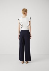 Hvit topp med svarte volanger, kombinert med høyhalsede marineblå vide bukser, med en glatt tekstur og minimalistisk design.