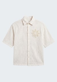 Chemise à manches courtes couleur crème, boutonnée, avec un motif géométrique texturé et une grande application fleurie sur la poitrine gauche.