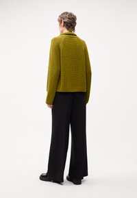 Giacca a maglia verde oliva con collo strutturato e maniche raglan, abbinata a pantaloni larghi neri e scarpe nere robuste.