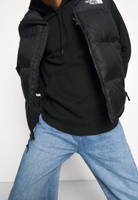 Un gilet matelassé noir sur un sweat à capuche noir, avec une fermeture éclair à l'avant et des poches. Un jean large bleu clair présente une texture délavée. Agrémenté de logos.