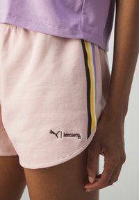 Gros plan d'une personne portant un short rose clair avec des bandes latérales noires, jaunes et violettes et un haut violet, la main relâchée le long du corps.