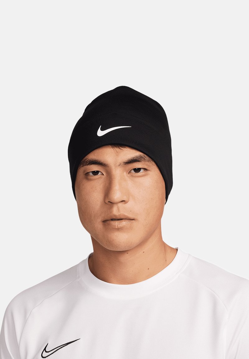 Beanie Nike nera con una texture morbida, con un logo bianco sul davanti. Indossato con una maglietta sportiva bianca.