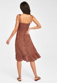 Robe florale marron avec des bretelles spaghetti, un corsage smocké et un ourlet volanté. Tissu léger avec un design fluide ; portée avec des sandales plates.