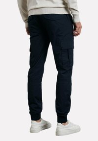 Pantalon cargo bleu marine avec poignets élastiques, plusieurs poches latérales et une texture lisse, associé à des baskets blanches sur un fond neutre.
