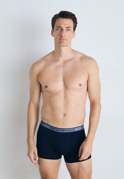 Fekete pamut boxer alsónadrág szürke elasztikus derékpánttal, melyen az "Emporio Armani" logo látható; a modellen meztelen törzs látható, semleges háttérrel.