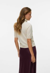 Kortebloezige licht beige gebreide trui met hoge kraag, gecombineerd met een lange, vloeiende donkerpaarse rok. Glad textuur, aansluitend op de taille.