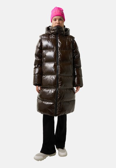Vrouw in een glanzende donkerbruine lange pufferjas, felroze beanie met tekst, zwarte broek en witte chunky sneakers, staande met haar gezicht naar voren.