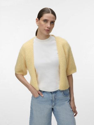 Vrouw met bruin haar in een staart, die een lichtgele korte mouwen cardigan draagt over een wit topje en lichtblauwe spijkerbroek, staand met één hand in de zak.