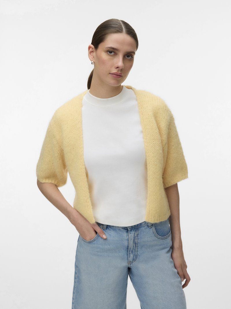 Femme aux cheveux bruns attachés, portant un cardigan jaune pâle à manches courtes par-dessus un top blanc et un jean bleu clair, debout avec une main en poche.