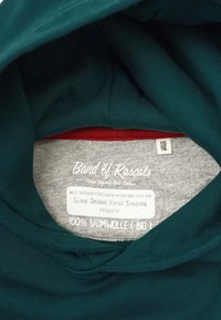 Tealfärgad hoodie med ett mjukt tyg, som har en grå etikett inuti. Etiketten säger "100% ekologisk bomull" och innehåller skötsel anvisningar och storleksdetaljer.