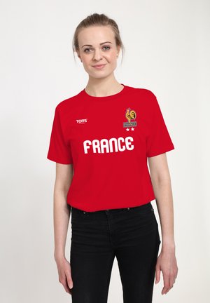 Ung kvinde iført en rød "Frankrig" T-shirt med en haneemblem, står og smiler mod en ensartet lys baggrund.
