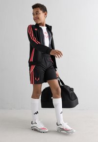 Boy in black and red sportswear carrying a black sports bag, wearing white socks and white cleats, standing against a plain white wall.