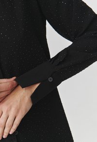 Black long-sleeve shirt featuring a textured dotted pattern and buttoned cuffs, made from a smooth fabric. Close-up of cuff detail shown.