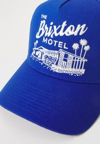 Blå tygkeps med vit broderad text "The Brixton Motel" och en grafik av ett motell med palmer.