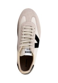 Zapatilla con una parte superior de cuero y ante beige, acento diagonal negro, cordones blancos y una etiqueta de marca en la lengüeta. La suela de goma es visible.