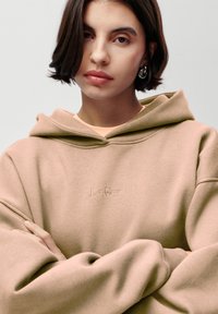 Felpa con cappuccio beige dal tessuto morbido, con logo ricamato "LeGer", maniche ampie e vestibilità rilassata. Il modello ha le braccia incrociate.