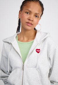 Graue Zip-Hoodie aus weichem Material, mit einem Patch im Design eines roten Herzens und Text, übereinander getragen mit einem hellgrünen gerippten Oberteil.