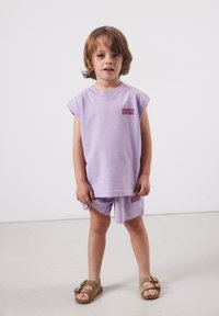 Jeune enfant debout, portant un débardeur violet clair assorti à un short avec le texte "American Vintage", et des sandales beige.