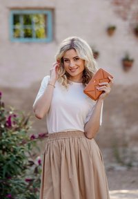 Beige, pleated skirt and white t-shirt. Orange wallet with geometric design held in the right hand. Green foliage and brick wall in background.