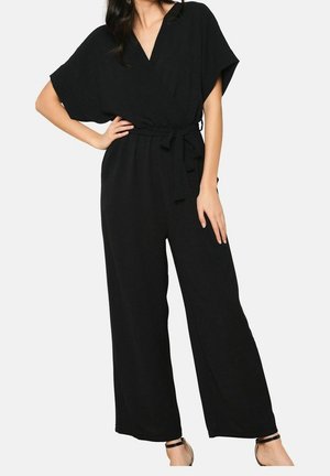 Kvinde iført en sort bredbenet jumpsuit med V-hals, korte ærmer, en knyttet taljebælte og sorte ankelremssandaletter.