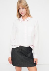 Mulher loira vestindo uma camisa de botão rosa claro, utilizada por dentro de uma saia mini cinza escura com bolsos laterais, de pé contra um fundo branco.