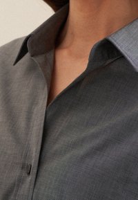 Närbild av en person som bär en oöppnad grå skjorta med krage, som visar halsen och övre delen av bröstet.