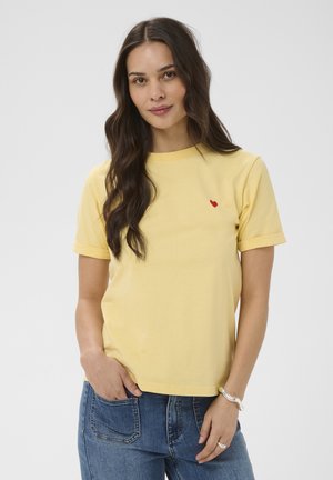 Žena s dlouhými hnědými vlasy, oblečená v žlutém tričku s malým červeným srdcem a modrých džínách, stojící s jednou rukou v kapse.