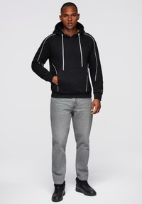 Junger Mann, der einen schwarzen Kapuzenpullover mit weißen Nähten, graue Jeans und schwarze Sneakers trägt, steht vor einem schlichten hellen Hintergrund.