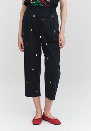 Pantalones negros recortados con pequeños estampados coloridos de frutas y mariposas, usados con bailarinas rojas, mostrando desde la cintura hasta los pies.